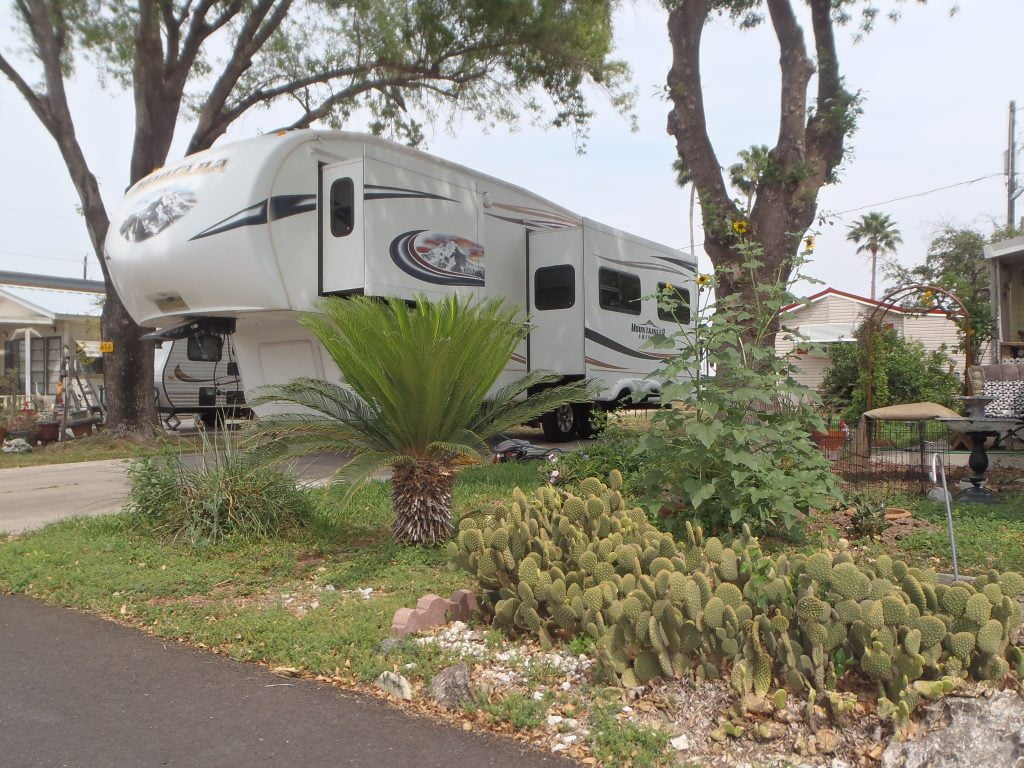 A photo of a Fifth Wheel camper parked on a treed lot, speckled with cactus and palms.