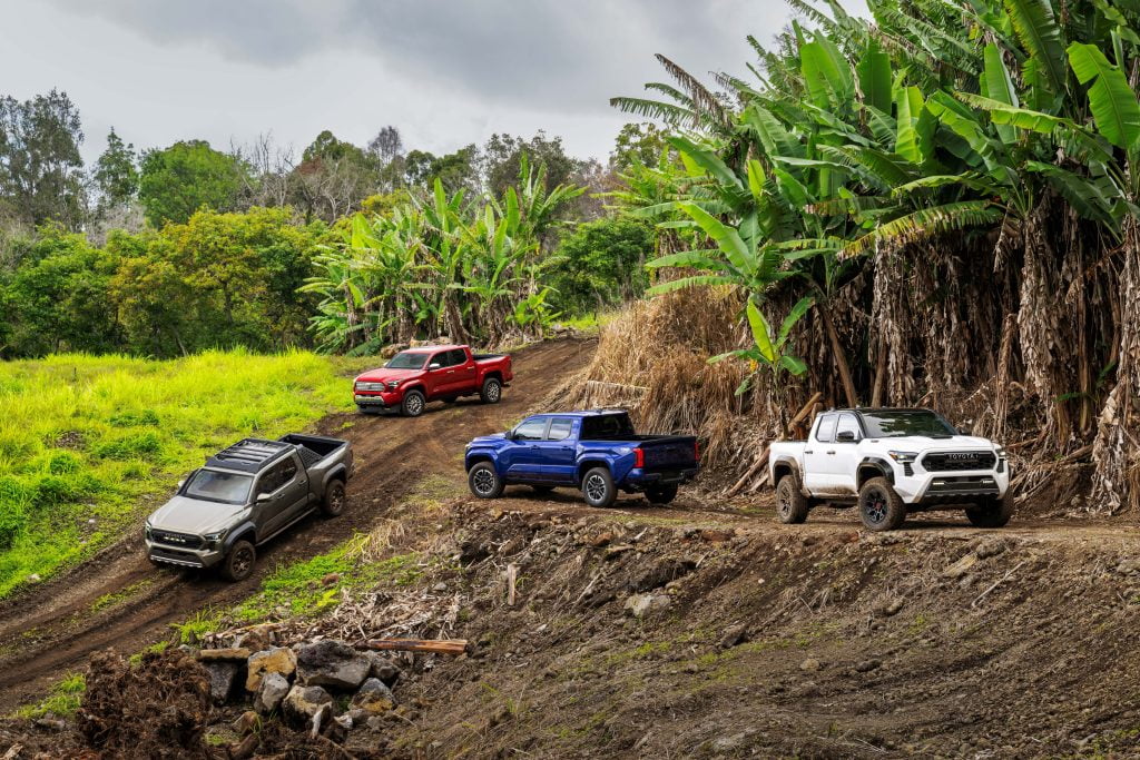 A picturesque scene in Hawaii featuring four Toyota 2024 trucks parked on a rugged landscape. In the beautiful lush green background is a white truck, a blue truck, a red truck, and a silver truck, showcasing a range of vibrant colours against the natural backdrop.
