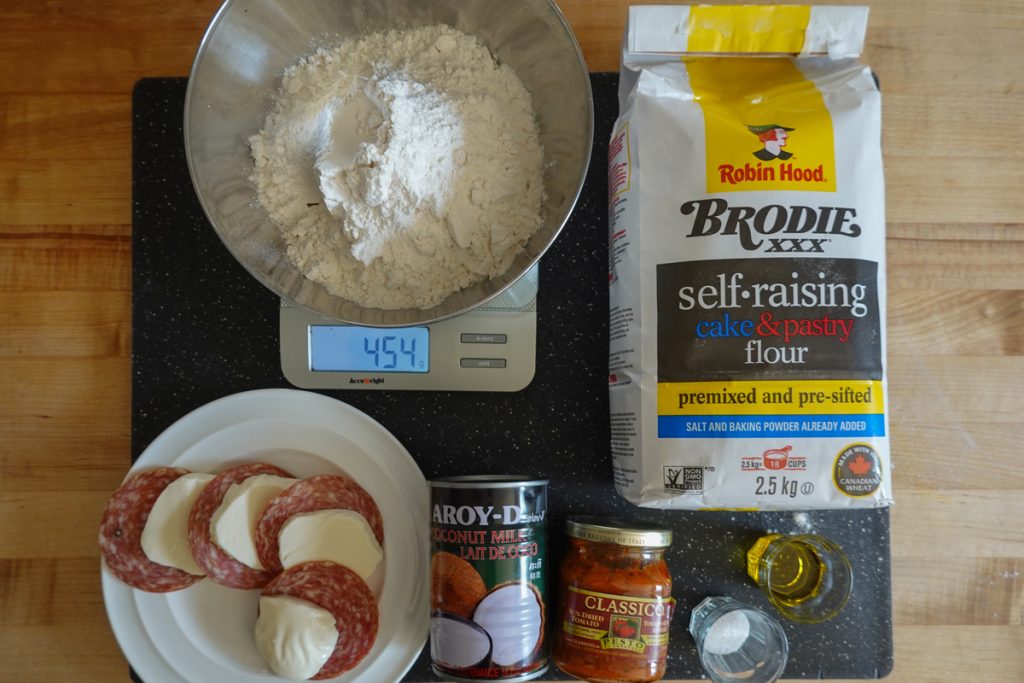 An image of ingredients used for the recipe. The image shows a bowl with flour, one bag of self-raising flour, olive oil, salt, tomato pesto, coconut milk, salami and mozzarella cheese.