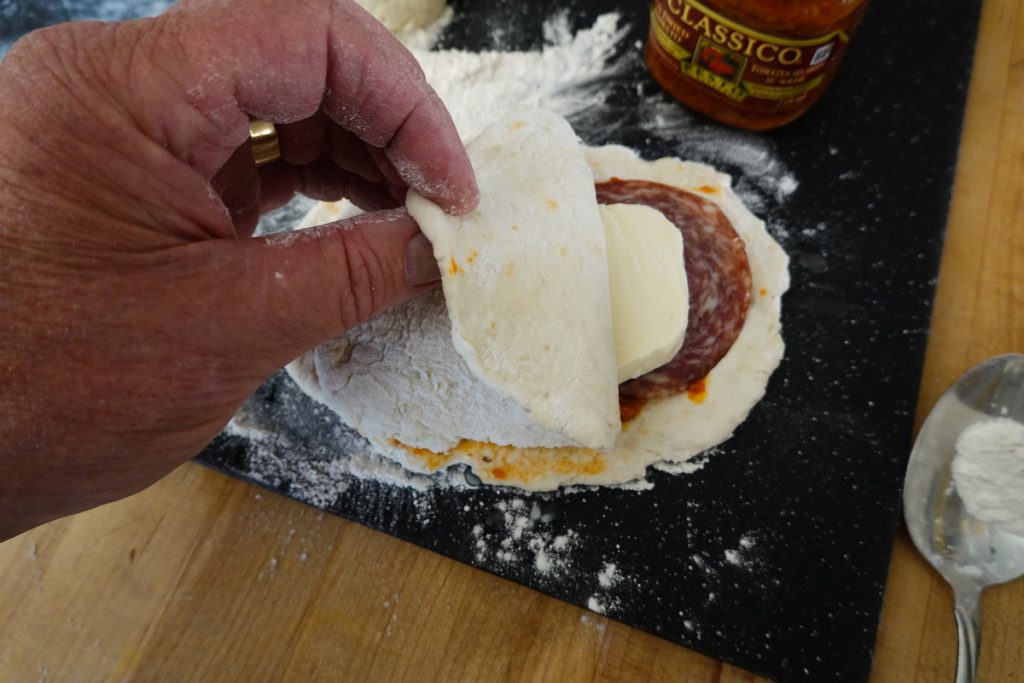 Close-up view of a man's hand skillfully folding the dough over a layer of salami, cheese, and sauce. The hands shape the ingredients into a mouth-watering filling, creating a delectable culinary masterpiece. The image captures assembling a delicious stuffed dough pie iron creation.