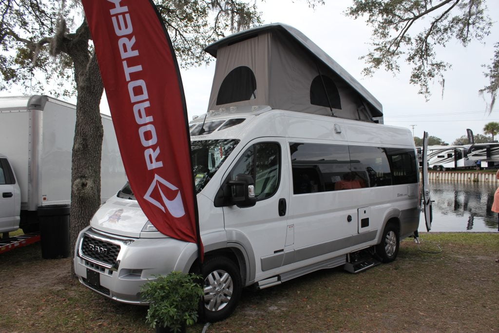 White Road Track camper van with an open rooftop tent. A red Road Track banner is displayed prominently on the side. Parked next to the van is a park truck near a tree. In the background, a scenic view of water can be seen, along with other camper vans in the distance.