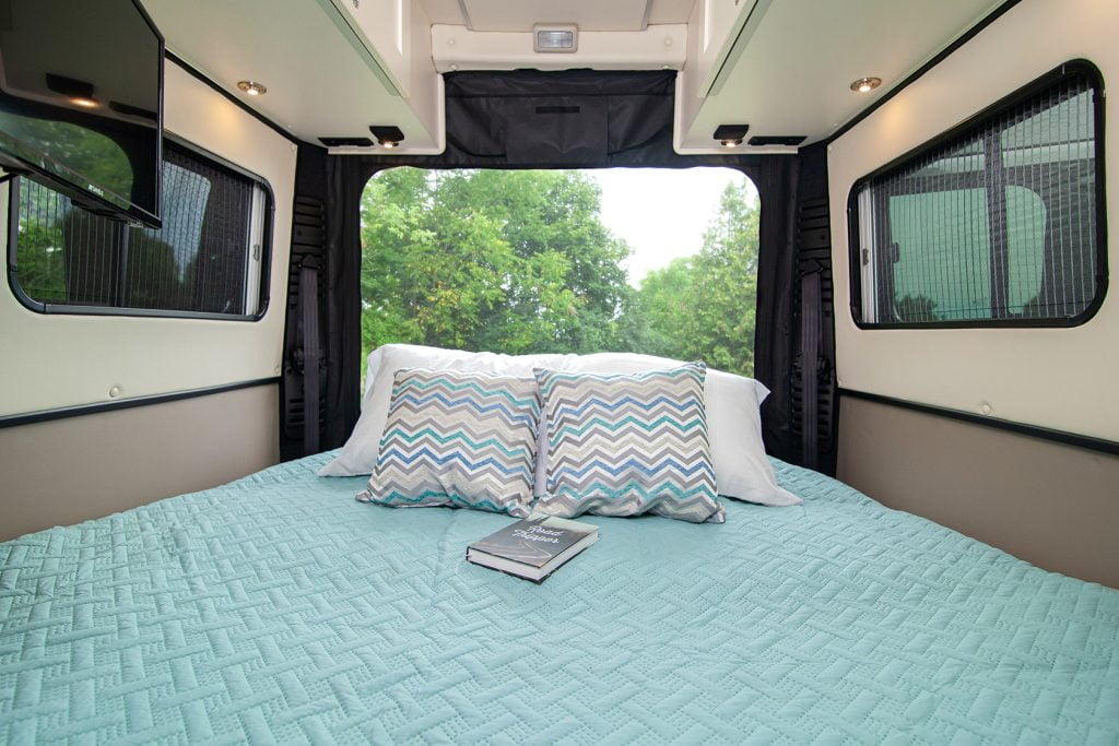 Interior view of a camper van featuring a comfortable bed, a TV in the corner, and two windows providing natural light. The back of the van is open, revealing a scenic backdrop of lush green trees.
