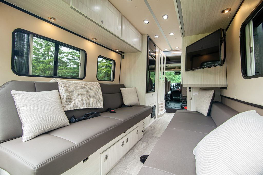 Interior of a Zion Slumber camper with cozy grey leather benches, tan pillows, and a blanket. A TV is situated in the corner, and lush trees can be seen outside through the windows, creating a serene atmosphere.