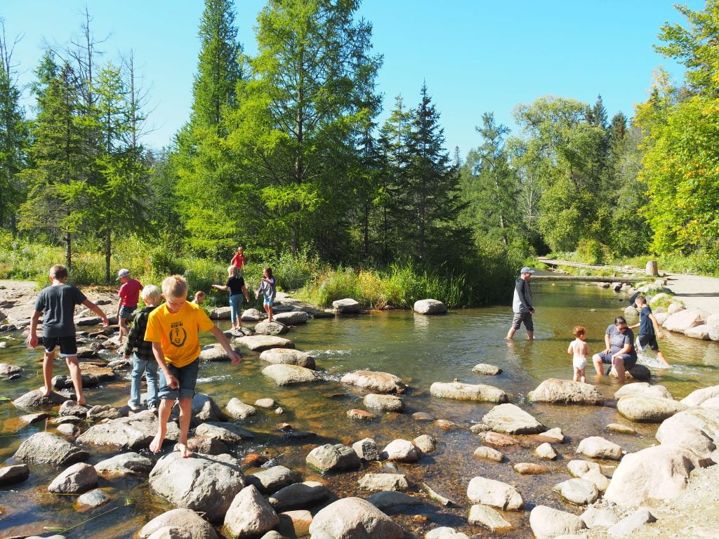 Groups of families walking on rocks, carefully crossing a shallow river. The riverbed is visible, with water flowing gently between the rocks. Lush trees form a backdrop, adding to the scene’s natural beauty. The image captures the sense of adventure and togetherness as families navigate the river, surrounded by the peaceful ambiance of nature.
