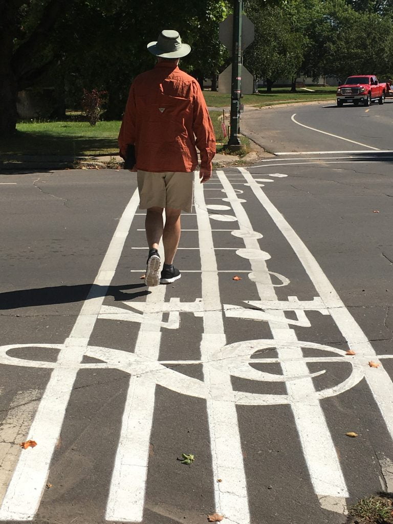 An image of a man wearing a rust colour jacket and hat is walking across a street painted with music notes.