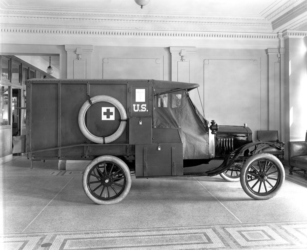 A black and white image of a 1917 Ford Model T Army ambulance displayed at the Ford Museum. The vehicle features the iconic Model T design with the Red Cross symbol on the side.