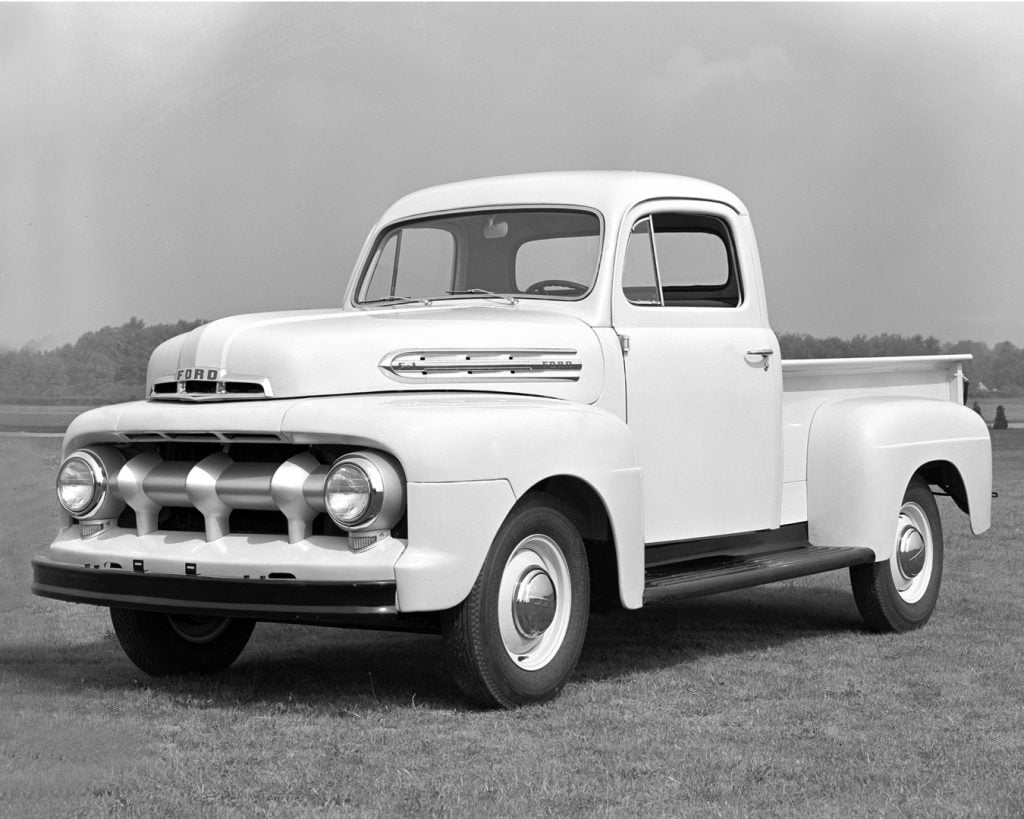 A black and white photograph of a white 1951 Ford F1 pickup truck, parked in a scenic location surrounded by trees. The vintage truck displays the distinct F1 design, capturing the essence of its era.