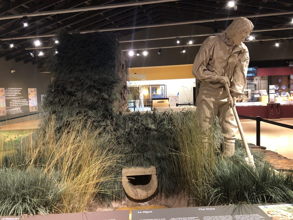 an image of an indoor museum photo featuring straw, and construction materials for a new straw house, and a concrete statue depicting a man engaged in work to create the building.