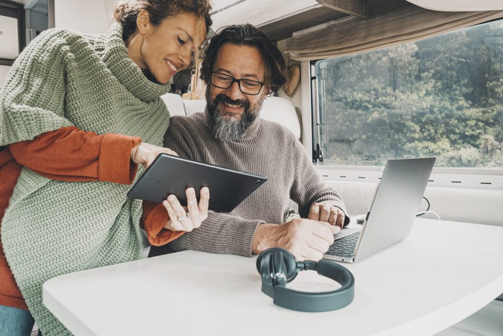 An image of a couple inside an RV parked in a wooded area. Both individuals are focused on a computer screen, engaged in an activity together.