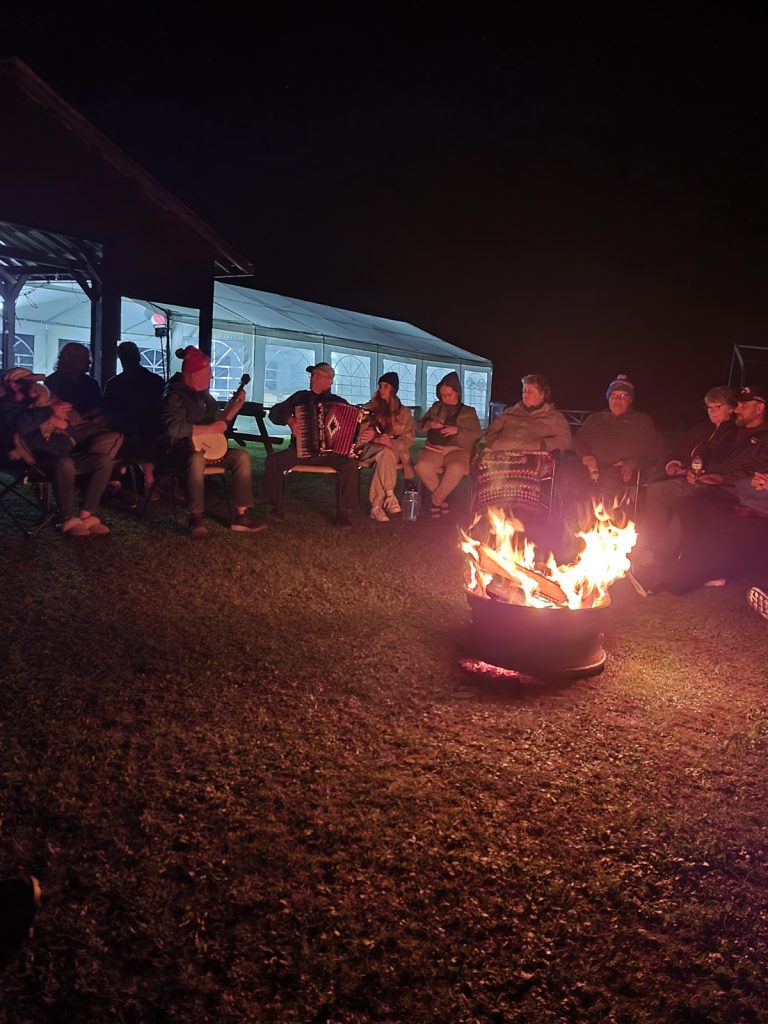 A crowd of people around a campfire at night. There is handclapping, an accordion player, and people appear to be singing.