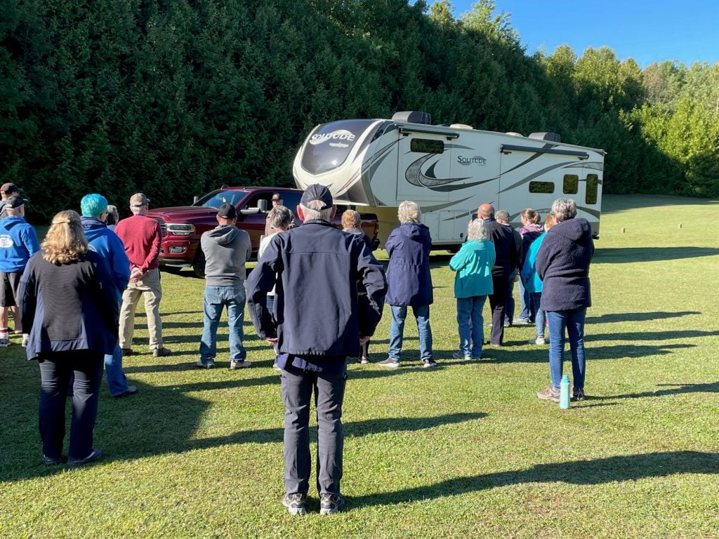 A group of people are gathered to watch an expert maneuvering his truck and trailer backing into a camping spot.