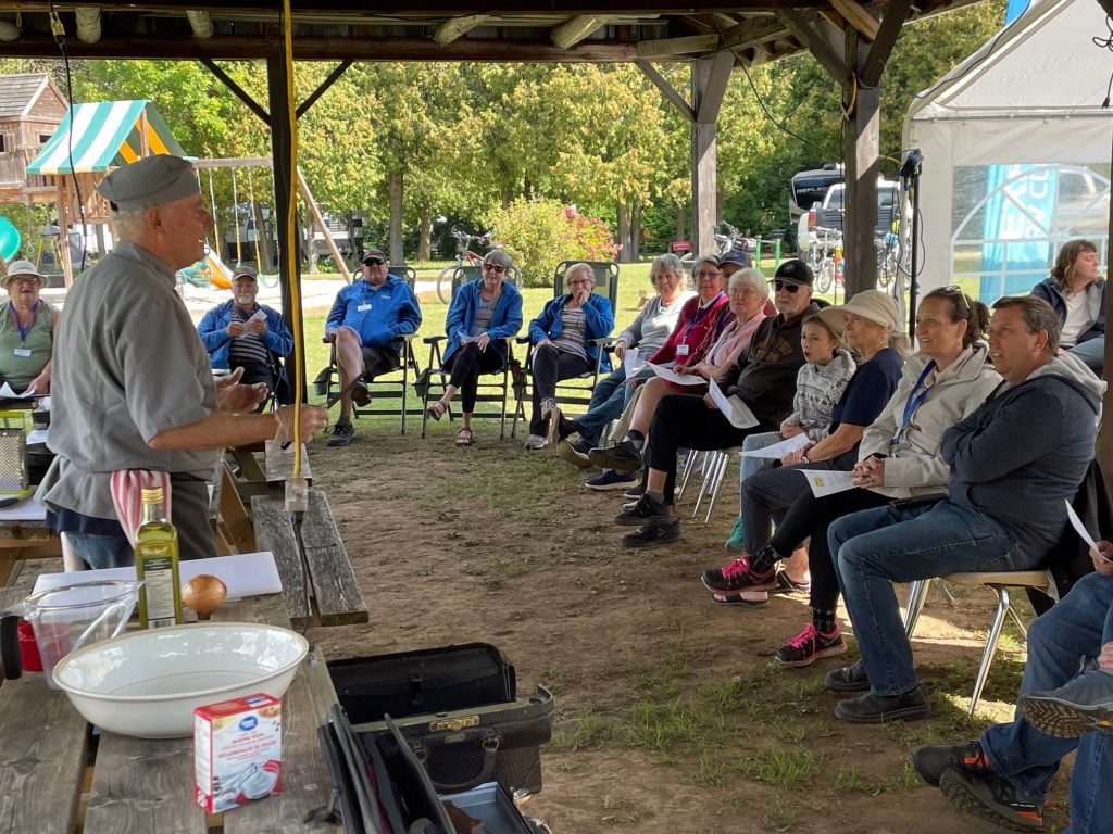 A Chef is giving a cooking presentation, on the table are delicious food ingredients. Around the Chef people are sitting in lawn chairs, engaged in the conversation.