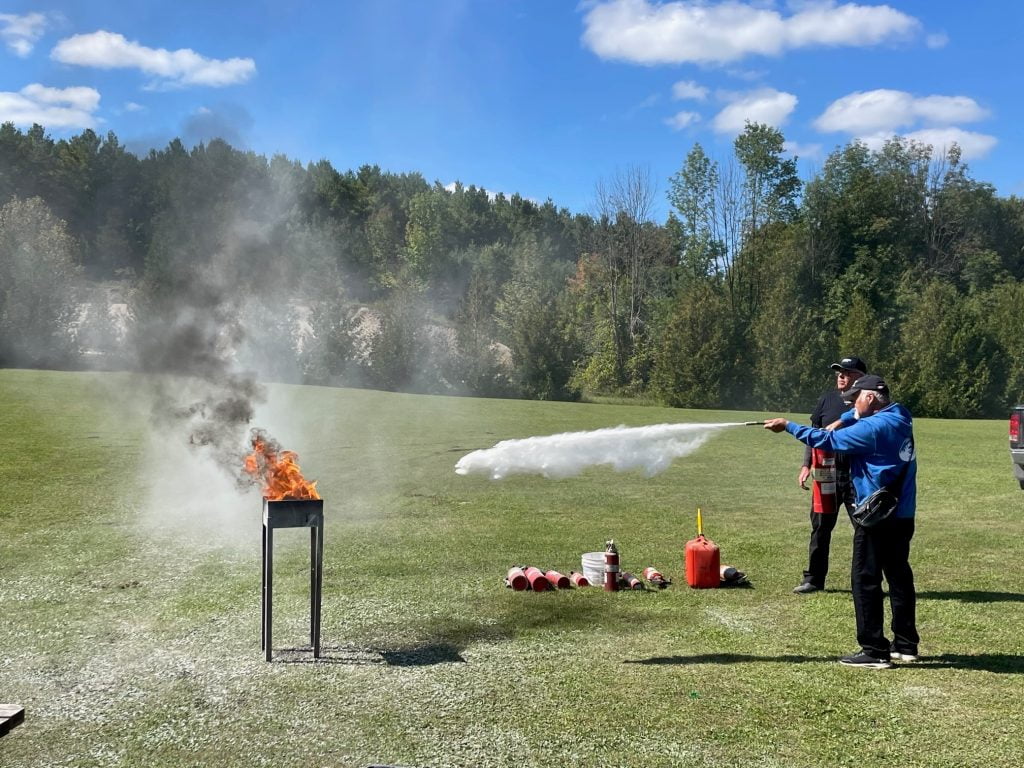 an image of a man using a fire extinguisher to put out a fire in a BBQ. A man is keeping watch in the background” On the ground are various cans and tanks used for the demonstration. In the background you can see beautiful lush trees.