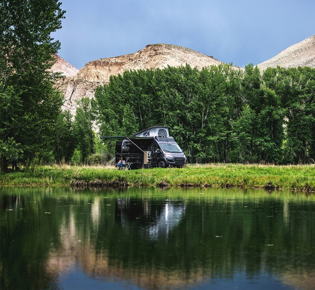 2024 Airstream Rangeline parked with an open awning, a woman seated underneath is relaxing and enjoying the RV lifestyle. She is surrounded by trees and towering cliffs in the background