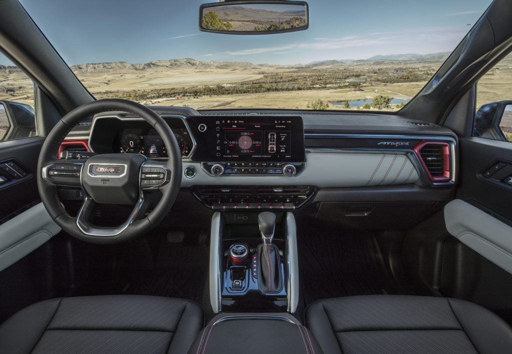 Interior view of a 2024 GMC Canyon featuring a modern dashboard with advanced technology. The steering wheel is on the left. Outside the window you can see sprawling plains with mountains beyond the plains.