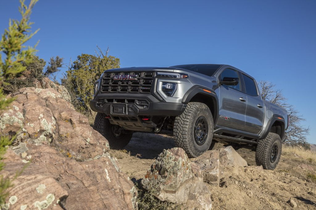 Silver 2024 GMC Canyon AT4X, four doors. Showing great ability with all terrain. Parked on a rocky incline with trees in the background.