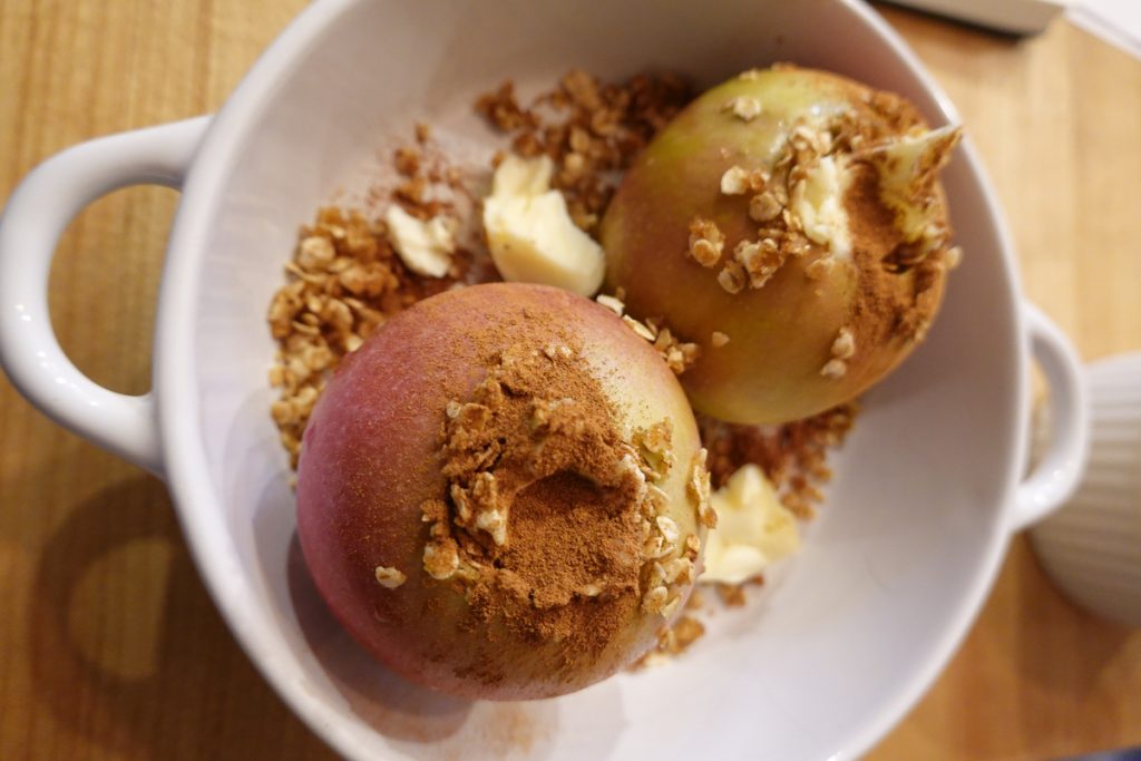 In a shallow bowl with two handles, you can see two apples that have been cored but not peeled. On top and around the apples you can see cinnamon and oats. The dish is ready to cook