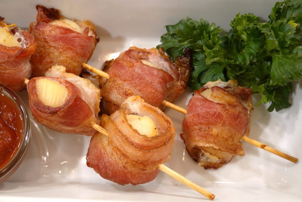 Perfectly cooked bacon is wrapped around scallops, pieced by skewers. There is bright green parsley on the plate to decorate the appetizer.