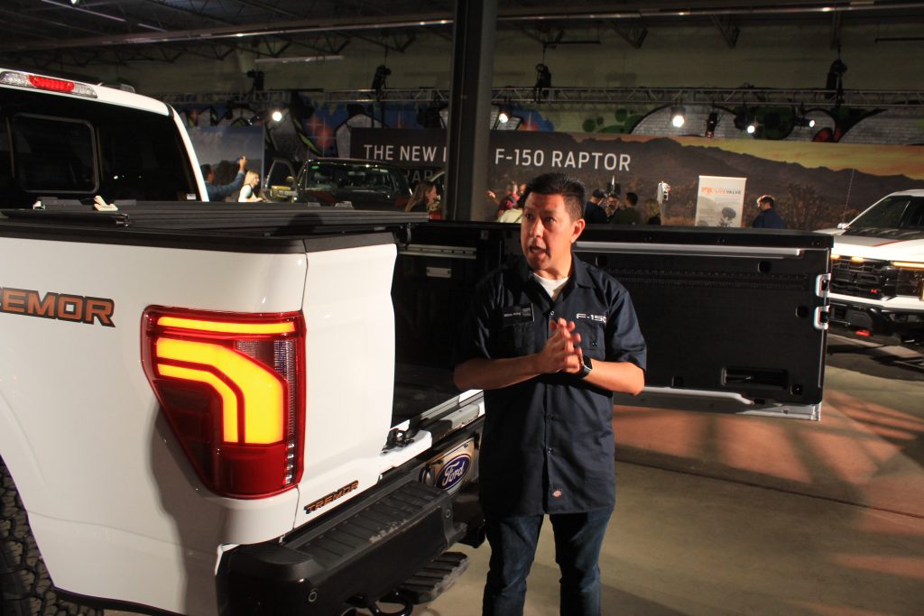 A while exterior F-150 with yellow and red lights can be seen close up. There is a man who appears to be giving a talk about the vehicle. Behind him, the door to the bed of the truck is open. The door swings out to the right instead of downward