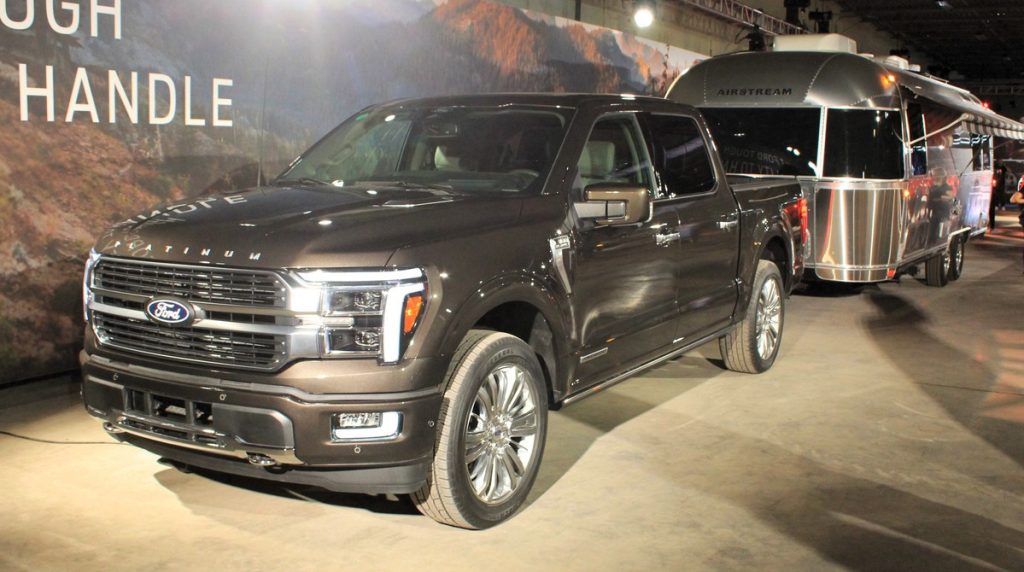 A silver four door Ford F-150 shines in the overhead lights, a sharp silver Air Stream is hitched to the truck. Behind the truck and the Air Stream is a back drop indicating an indoor trade show.