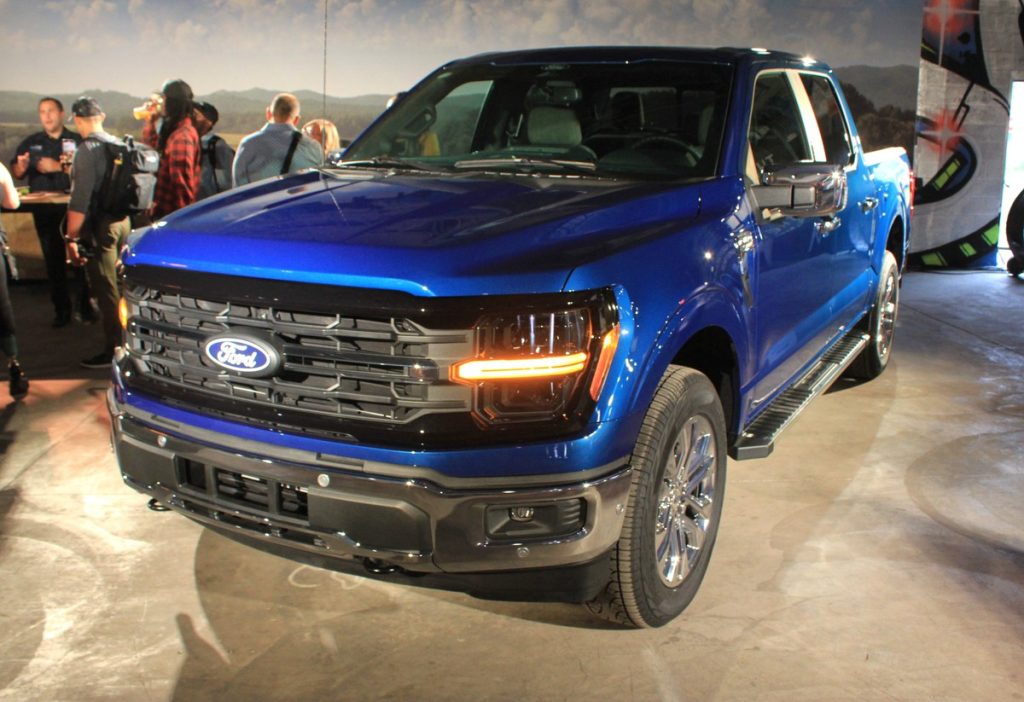 A large blue Ford truck is parked in a display area. In the background people can be seen mulling about and talking
