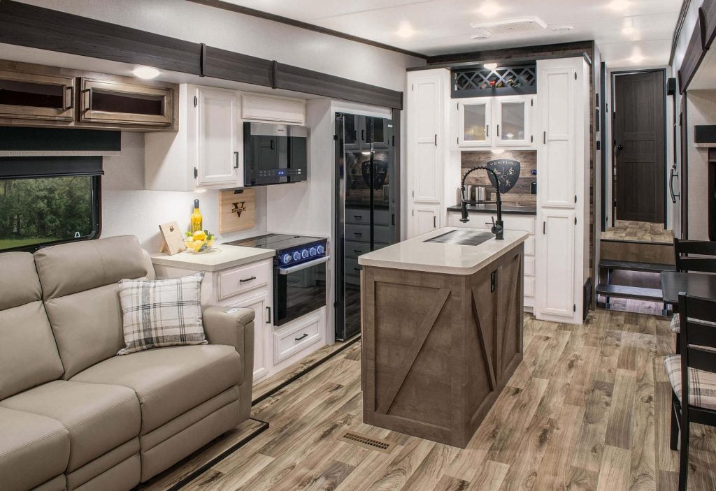 This open concept RV is like a home away from home. The leather sofa has lots of space in front of it. There is an island with a sink, a range with an oven and an overhead microwave. There is a full-size fridge in stunning black. There is a partial view of tables and chairs. In the rear there are steps leading to a wood door