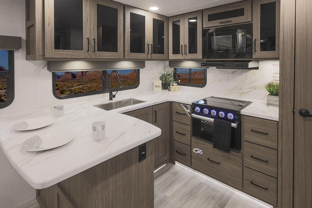 A gorgeous kitchen inside the RV. The modern kitchen has plenty of white counter space, a sink, and a built-in stove top. Above the stove is a build in microwave. There are lots of cupboards in natural wood shades.