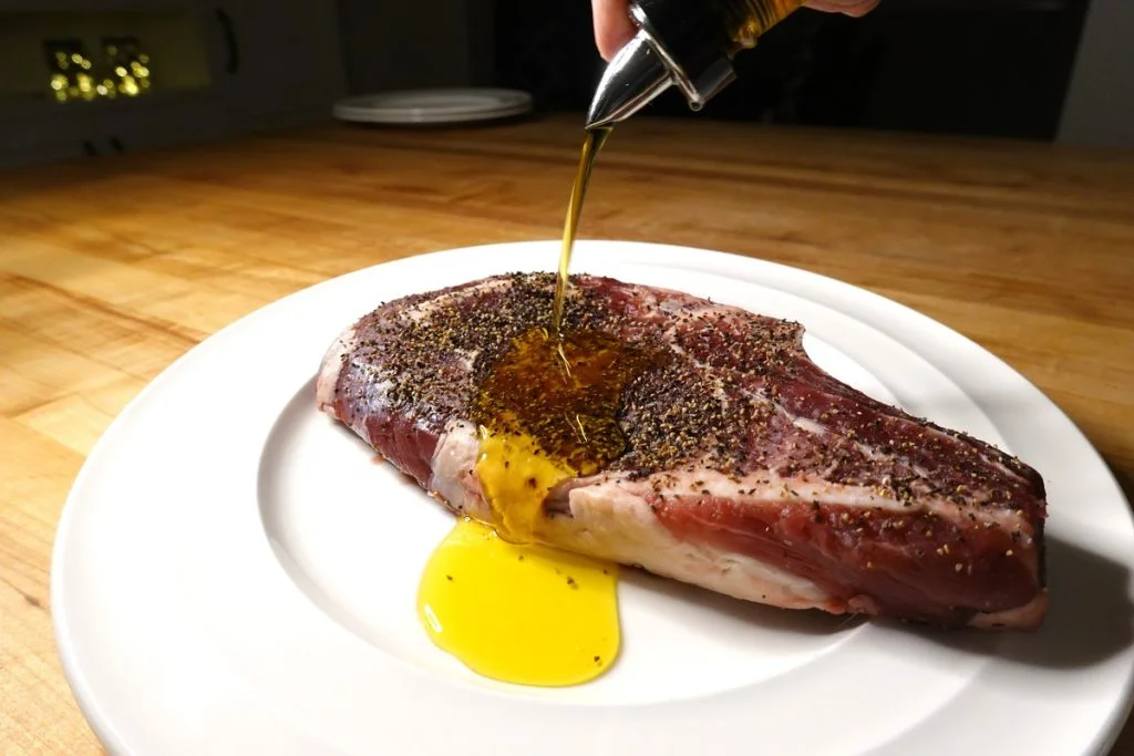 Olive oil poured over a seasoned steak on a white plate sitting on a wooden table.