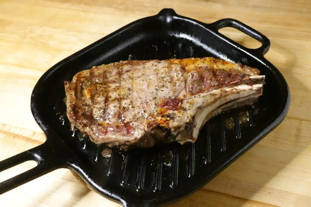 Cooked rib steak on a grilling pan cooled down and sitting on a wood counter.