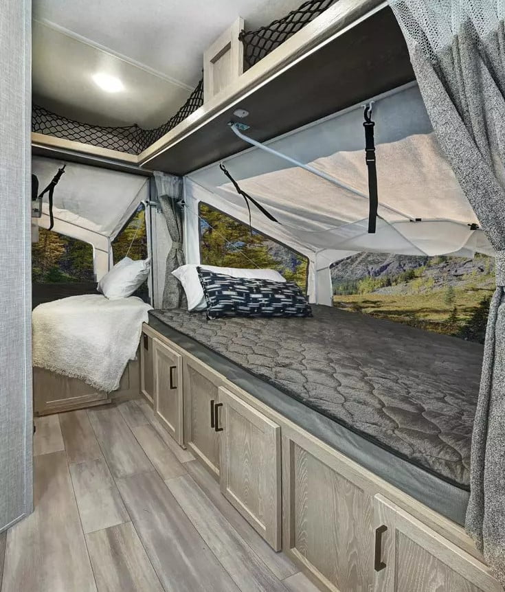 A beautiful wood floor and cupboards can be seen next to a pop out bed with a screen, allowing for fresh air and sunlight to stream through the RV.