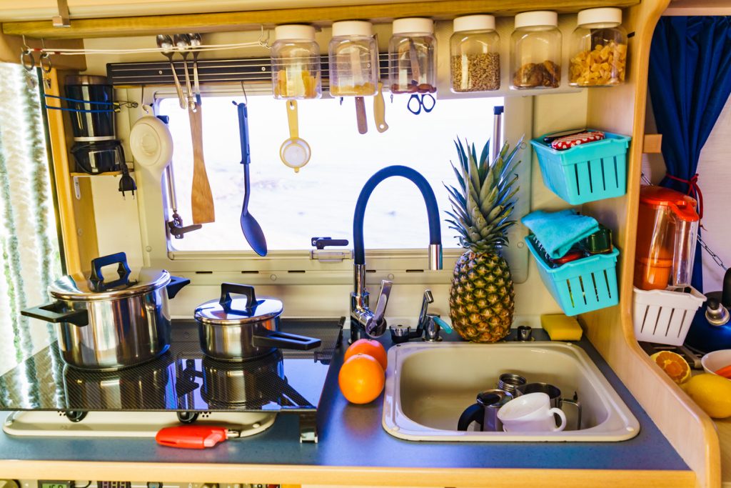 Close up picture of a kitchenette in an RV with pots on the stove and containers of different foods hanging from above sink and to the side suspended from the wall.