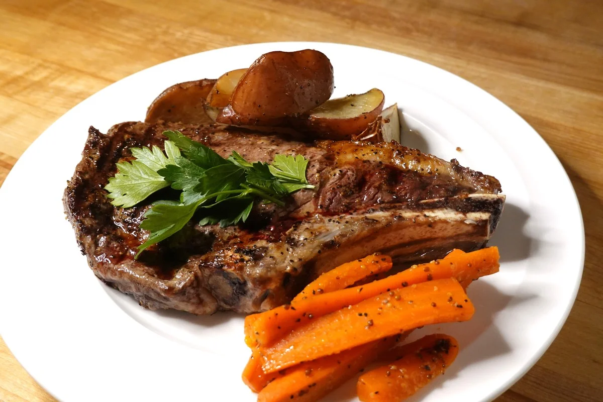Grilled steak garnished cilantro served with potato wedges and carrots on a white plate on top of a wooden table.