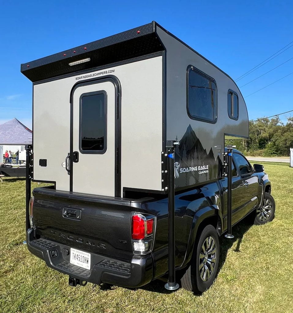 A black pick up truck has a traditional looking camper attached. The camper is beige with black accents. In the background you can see trees.