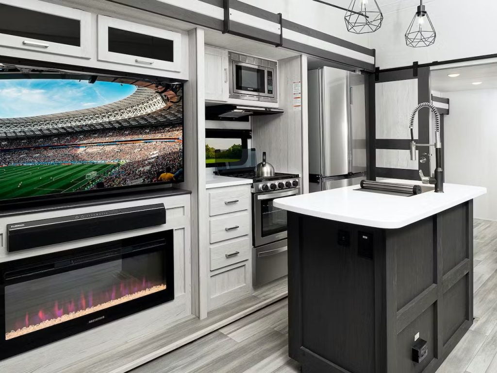 This RV showcases a television above a fireplace with storage above the television. There is an island with a built-in sink in front of a stove and oven. Above the island are two stylish hanging lights. There are three drawers for storage.