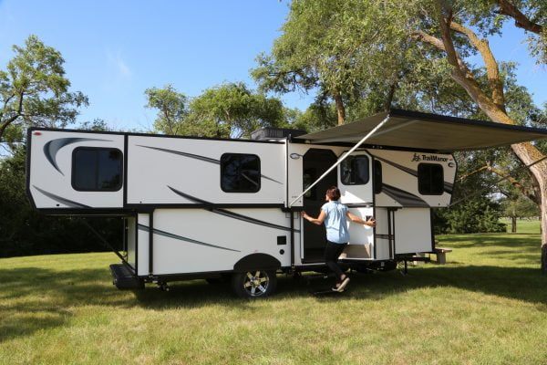 An image of a TrailManor Sport 2720QB. The travel trailer is parked on a grassy area. There are trees in the background and to the right of the trailer. The trailer is white with a black awning. There are three windows. The door is open and a young boy is going into the open door.” width=