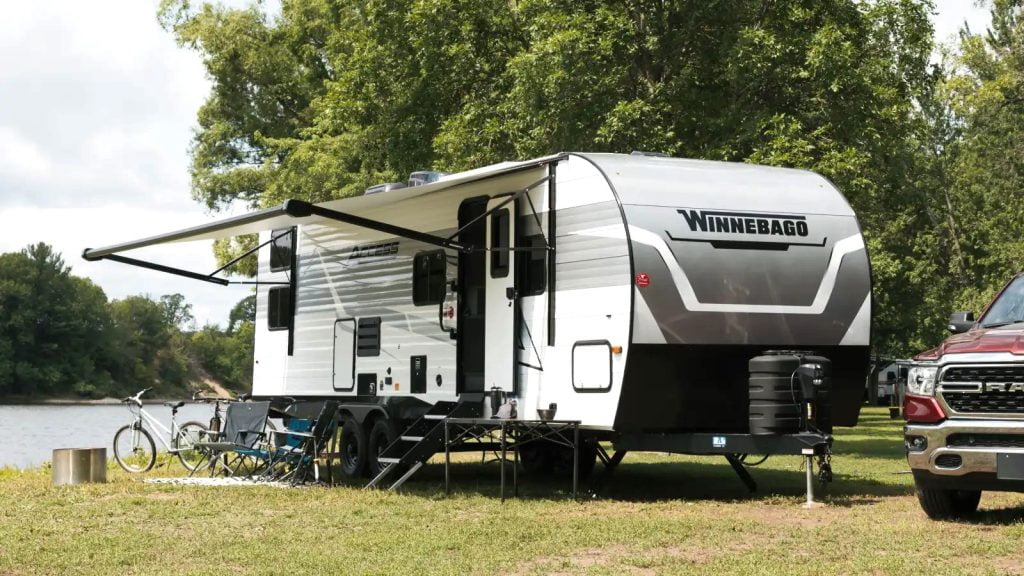 A Winnebago Access is parked next to a lake. There is a red truck partially seen beside the trailer. The trailer is white with grey accents. The awning is open blocking the bright sunlight. There are trees in the background