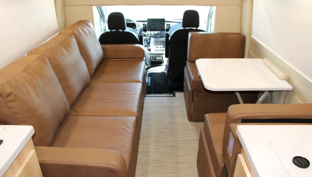 An image of a class B interior. There is a brown leather sofa. Across from the sofa is a small dining table and seats for two.