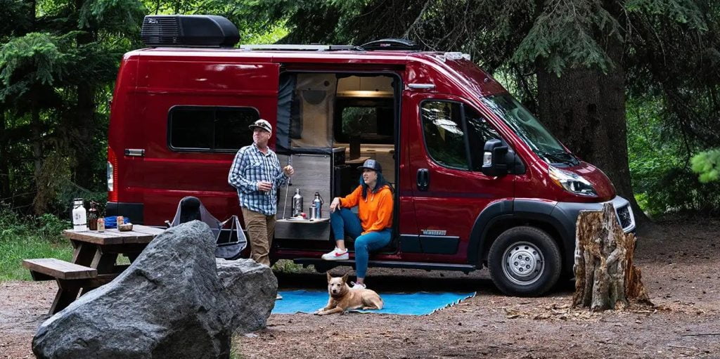 An image of a couple with an adorable dog are enjoying a meal outside of their ruby red Winnebago Solis Pocket.