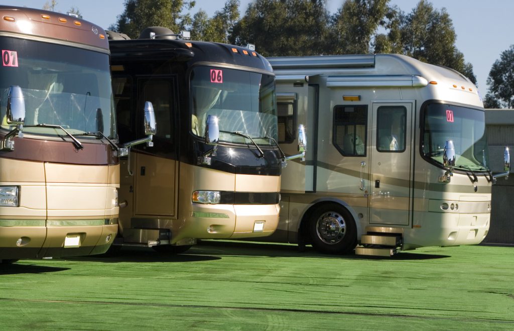 A stunning view of three gorgeous RV’s parked next to each other” Each RV is as captivating as the next. It’s a sunny bright day and the sun is reflected off of the pristine windows. The RV’s are parked on green grass.
