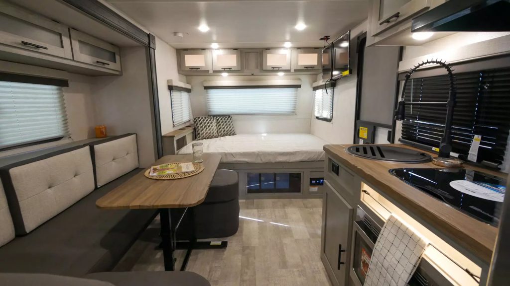 An image of a sleek interior of an RV. Closed blinds sit in front of a black stove and sink with a sink cover. A striped towel is hanging on the handle of the oven. On the left is a small table and bench. In the rear of the RV is a small bed.