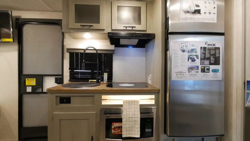 An image of an RV kitchen. A stainless-steel fridge sits on the right and a small sink and stove is in the centre. There is a door next to the sink and stove