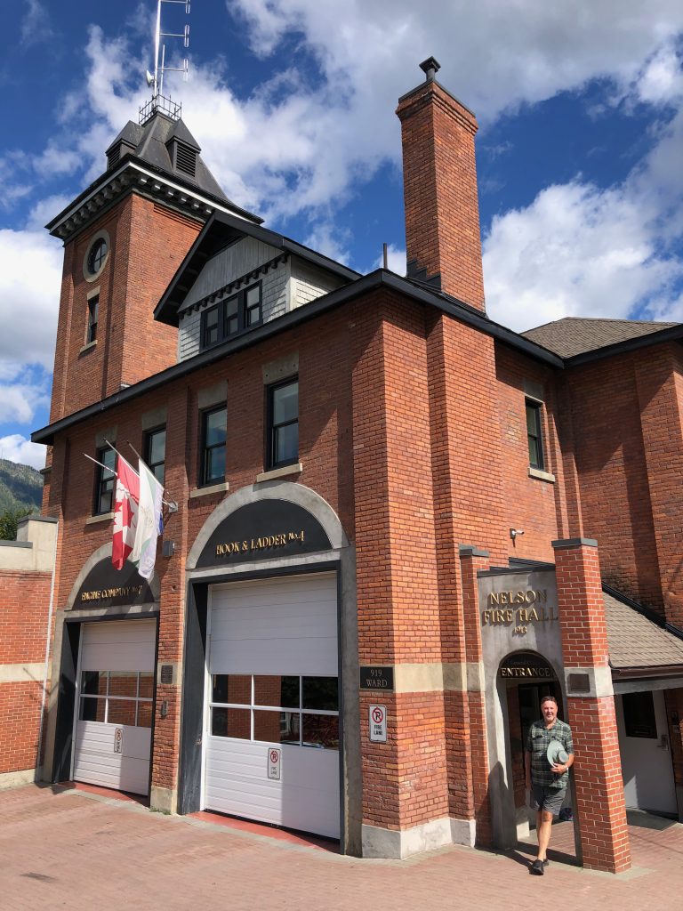 A man standing in front of the Nelson Fire Hall that has red brick and two garage doors. 