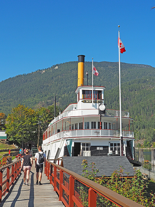 Two people walking on a dock next to the S.S Moyie in Kaslo with a view of the forest landscape behind it. 