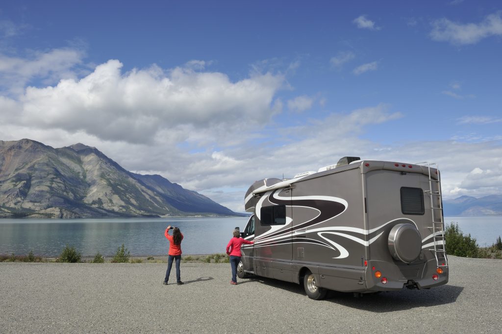 An image of an RV parked in a parking lot. There is one person in a red shirt and denim jeans who is next to the RV. The other person is taking a photo of the stunning view of the lake with mountains in the background.