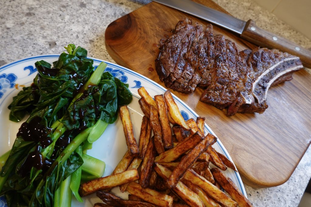 Gai lan and French fries on a plate with a cooked Rib steak on a cutting board next to it ready to be served.