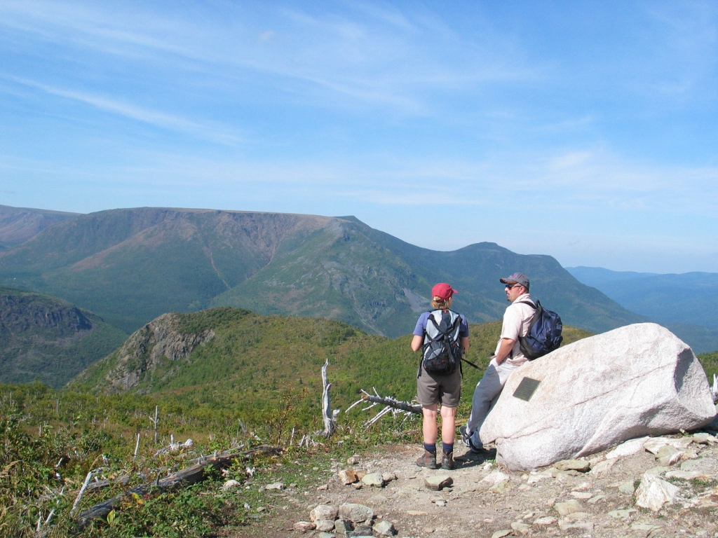 A man and a woman stand next to a rock with the Appalachian Mountains in the background at the G Parc National de la Gaspesie.