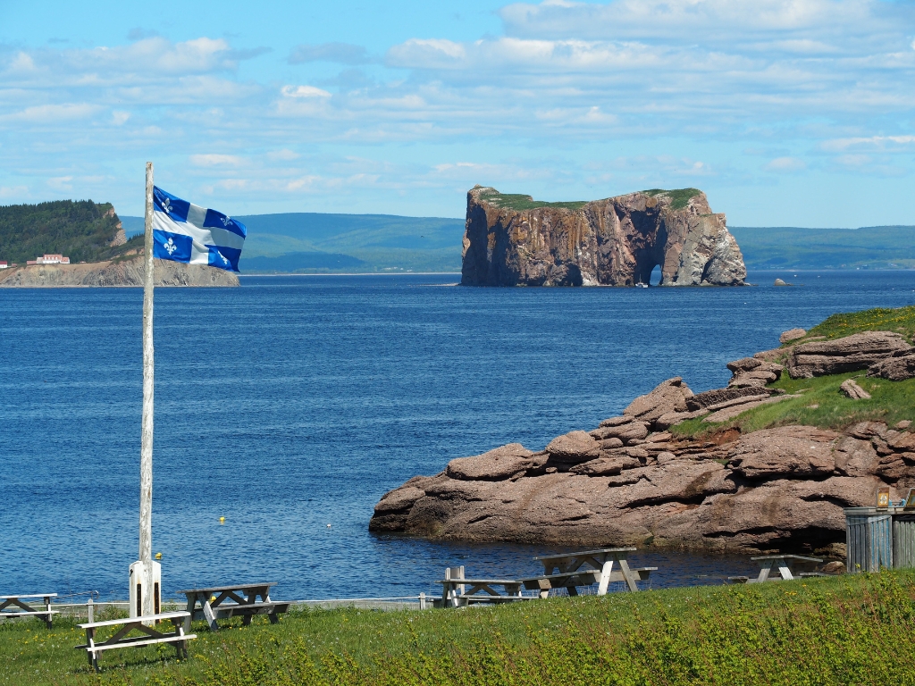 View of eye-in-the-needle Percé Rock, a massive piece of limestone cracked from the coast and sculpted over time by the sea and the wind, a Quebec flag flies in the wind.