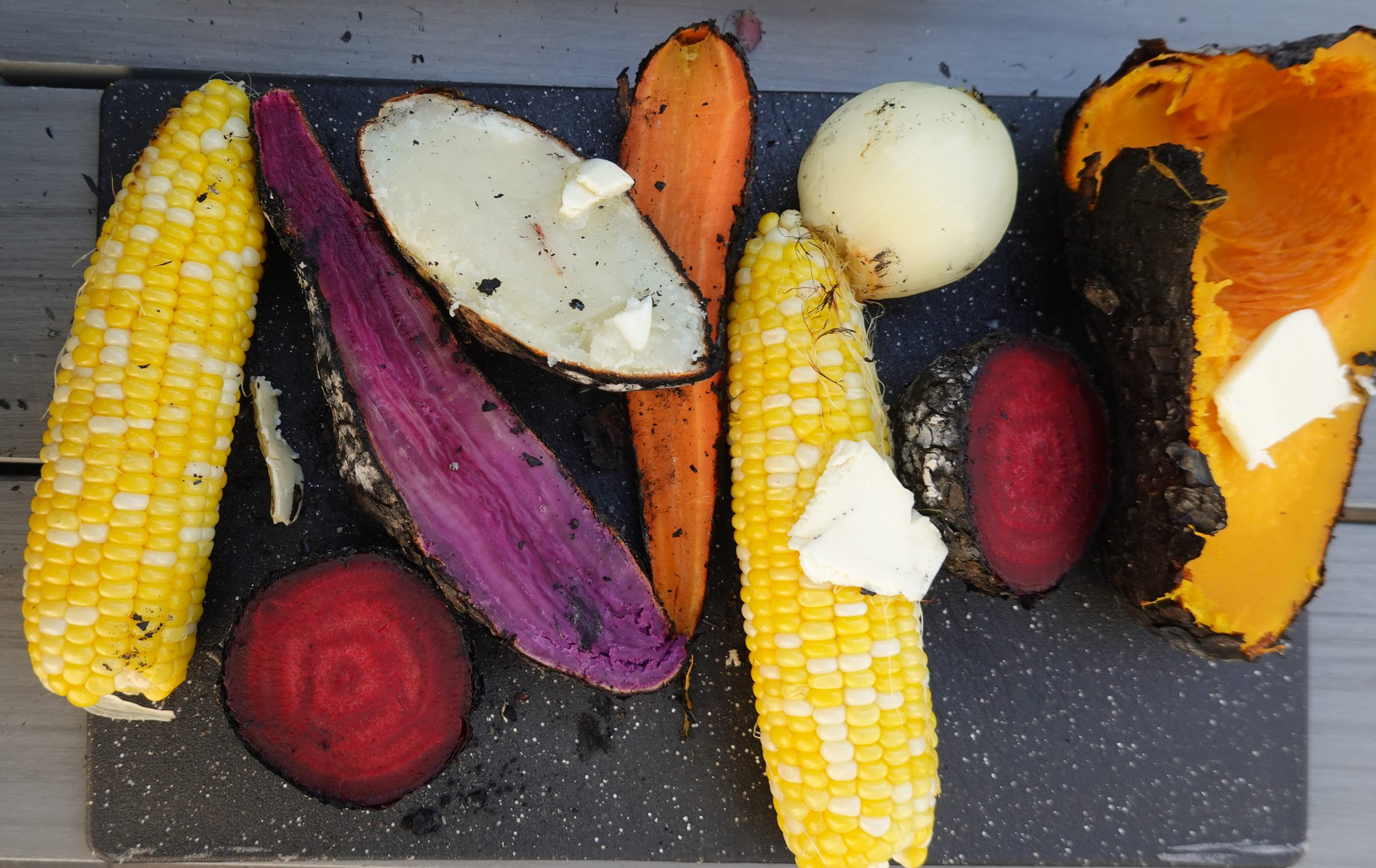 Image of cooked vegetables on a cutting board. From left to right roasted corn, beet, purple yam, russet potato, carrot onion and squash.