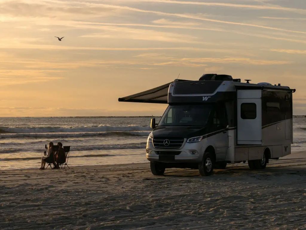 A couple sitting in chairs at the beach watching the sunset in front of their Winnebago View parked in the sand.