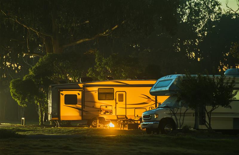 Motorhomes and Fifth Wheel Travel Trailer are parked at a campsite surrounded by trees at dusk. The RVs are both lit by the last rays of the setting sun, casting long shadows across the campsite. The scene suggests a peaceful and relaxing camping experience.